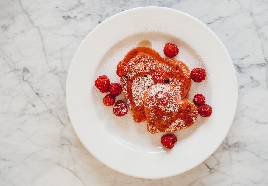 valentine's day breakfast idea with a white marble table with a white plate with pink heart shaped pancakes topped with raspberries, powdered sugar, and maple syrup