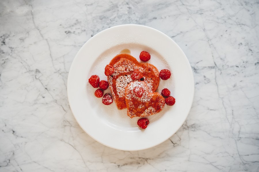 a white marble table with a white plate with pink heart shaped pancakes topped with raspberries, powdered sugar, and maple syrup