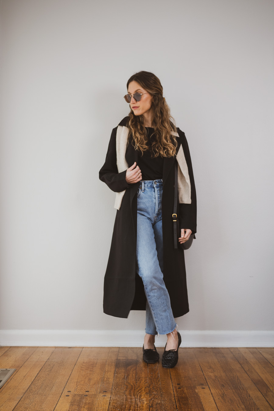 outfit with a black trench coat, black tee, light wash jeans, and black woven flats with a beige sweater worn over the shoulders