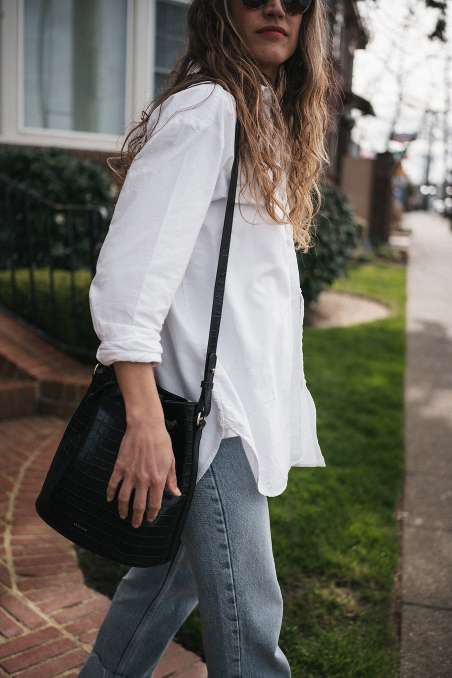 ashley wearing the Sézane Max Shirt in white, light wash cuffed jeans, black fisherman sandals, and round tortoiseshell frame sunglasses and a black crocodile print bucket bag