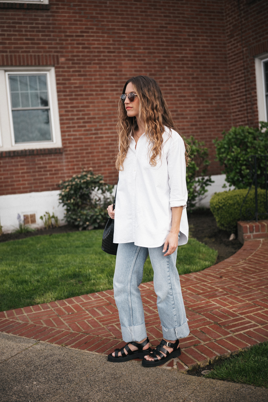 ashley wearing the Sézane Max Shirt in white, light wash cuffed jeans, black fisherman sandals, and round tortoiseshell frame sunglasses and a black crocodile print bucket bag