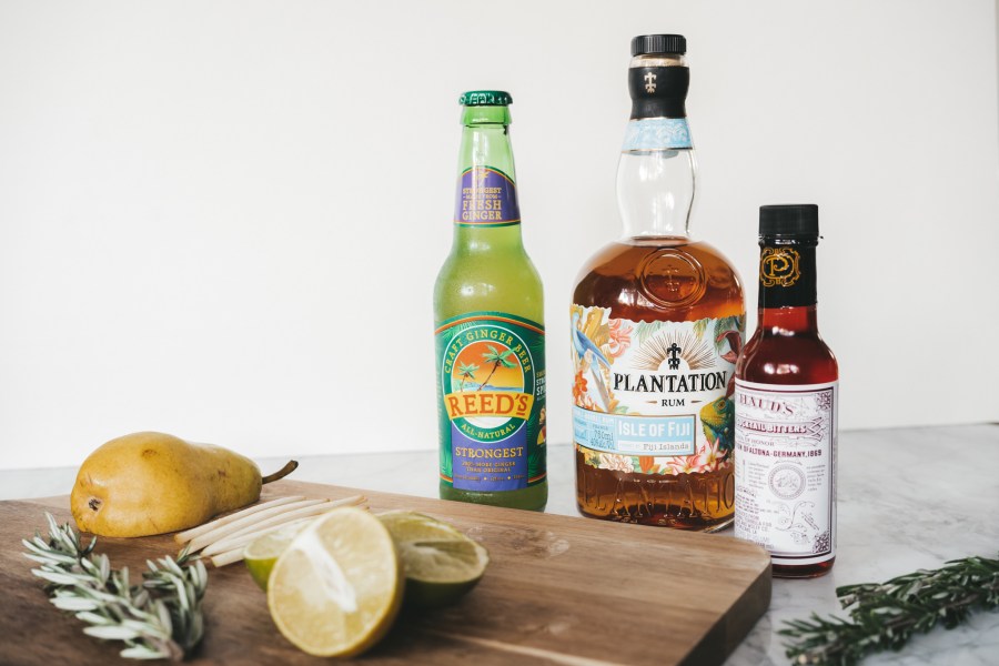 a bottle of ginger beer, a bottle of rum, a bottle of bitters, and a cutting board with a cut pear, sprig of rosemary and sliced lime