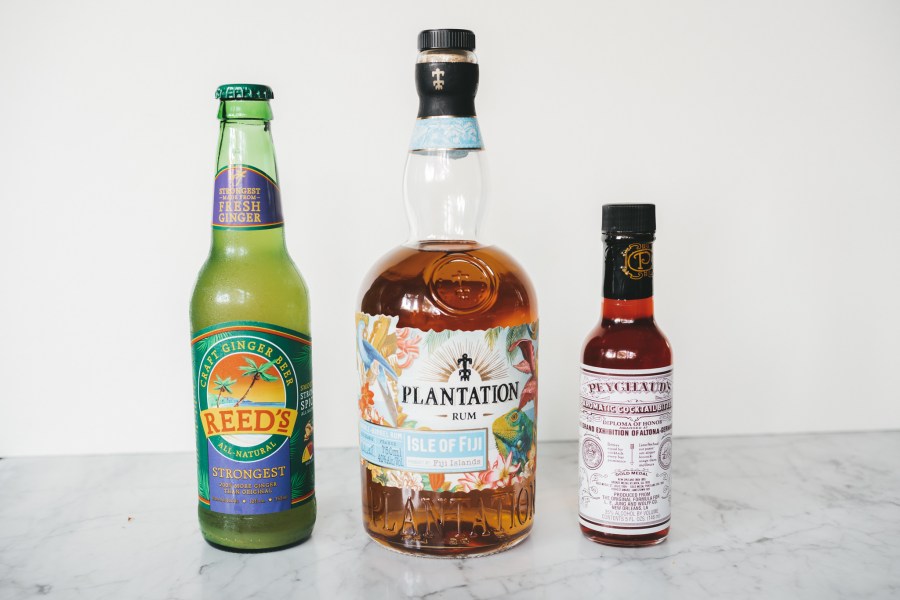 a bottle of ginger beer, a bottle of rum, and a bottle of bitters on a white marble table with a white background