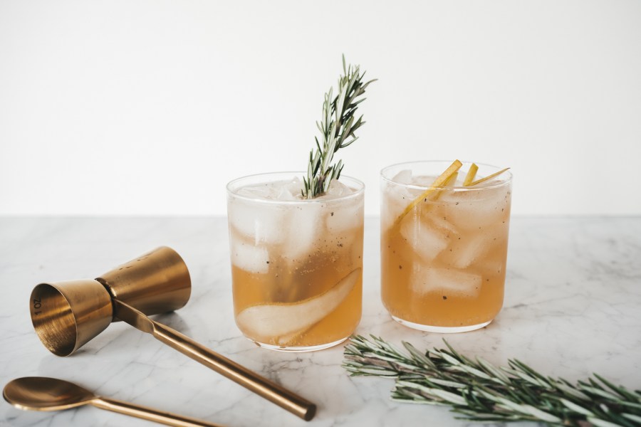 a picture of a rocks glass with a fall cocktail with pear and rosemary, a gold cocktail stirrer and a sprig of rosemary on a marble table