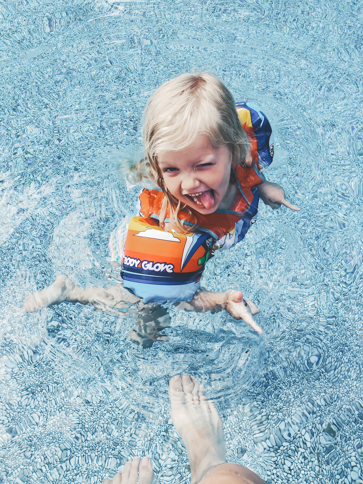 a little girl with a puddle jumper in a pool winking and smiling