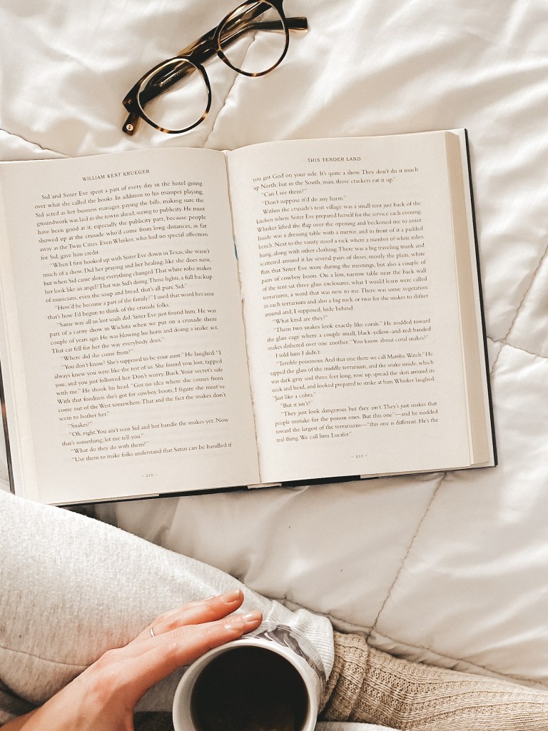 a book open on a white comforter, a pair of glasses, a woman's leg with gray pants and a taupe socks holding a mug tea