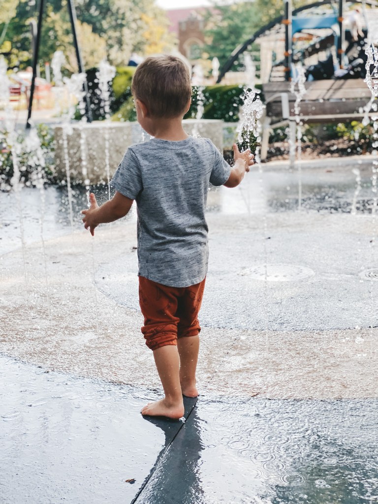 a toddler boy in orange pants pushed up to his knees and a gray t-shirt standing next to a sprayground