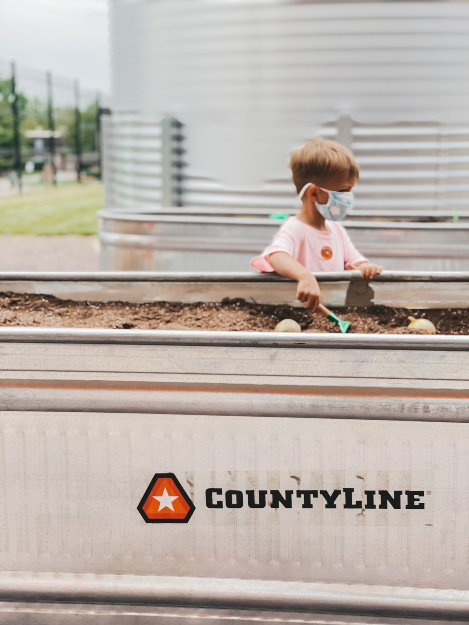 a little boy in a pink shirt standing next to a large metal tub with dirt and shovels in the outdoor exhibit at Wonderscope