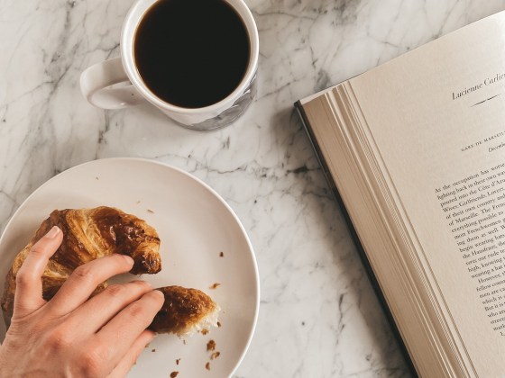 a marble table, a white plate with a croissant, a mug of coffee, an open book and a succulent