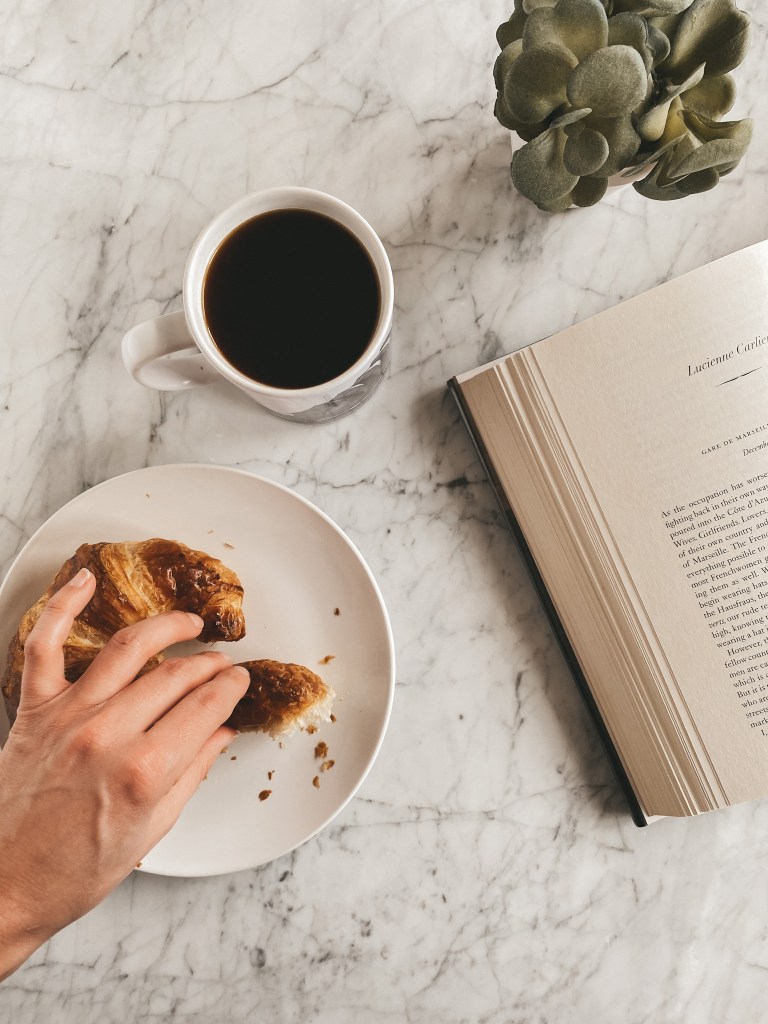 a marble table, a white plate with a croissant, a mug of coffee, an open book and a succulent