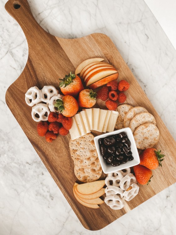 a wood serving board with yogurt covered pretzels, strawberries, sliced apples, crackers, white cheddar cheese, raspberries, and chocolate covered strawberries